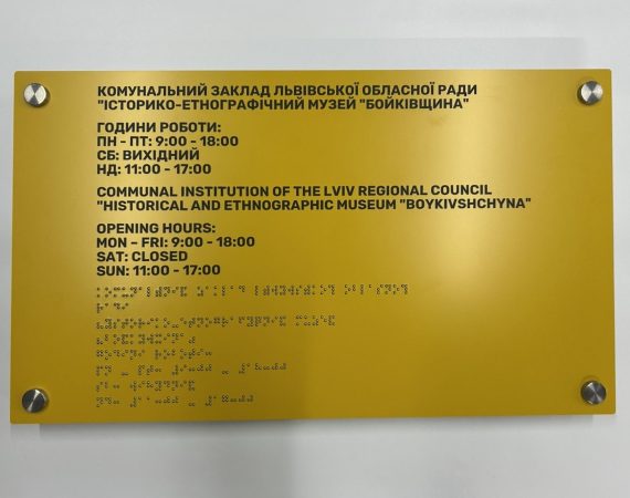Жовта інформаційна табличка музею «Бойківщина» з графіком роботи та текстом шрифтом Брайля