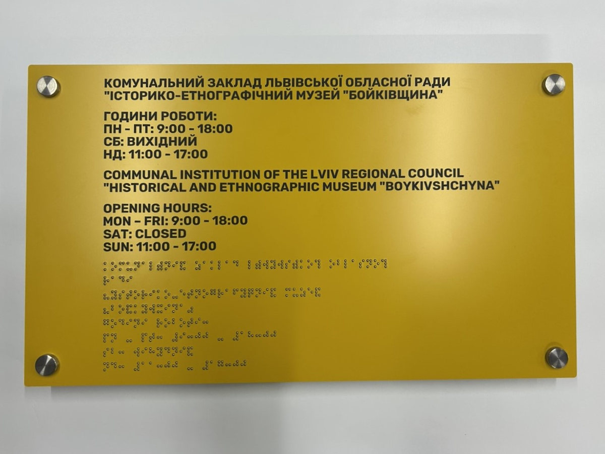 Жовта інформаційна табличка музею «Бойківщина» з графіком роботи та текстом шрифтом Брайля