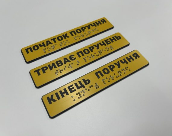 Три жовті таблички з написами «Початок поручня», «Триває поручень» і «Кінець поручня» та шрифтом Брайля